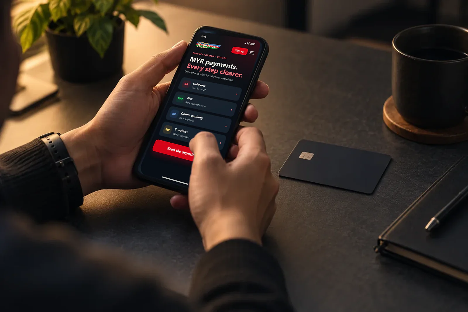 Hands holding a phone showing the 100cuci MYR payment guide, beside a bank card